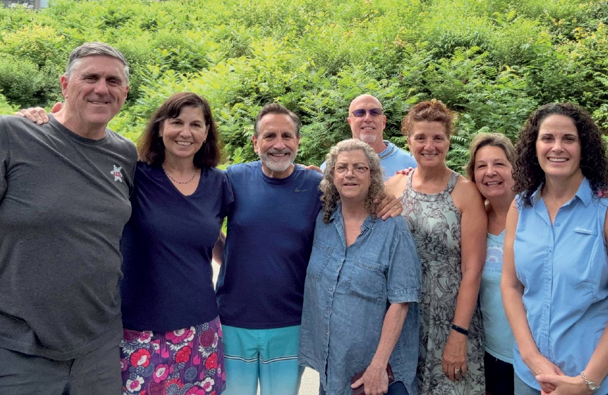 A group portrait of eight people standing outdoors in front of lush green trees and foliage.