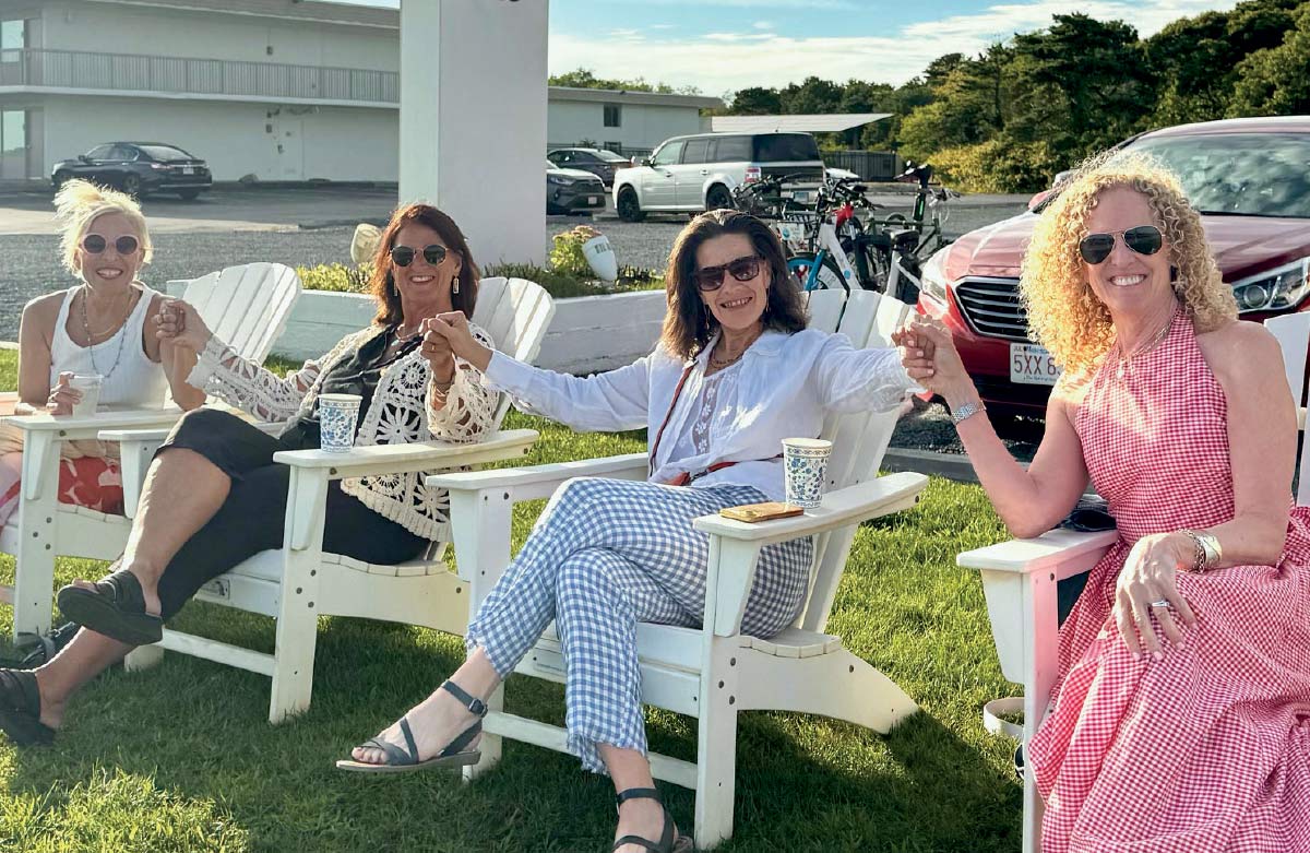 Four women sitting in white Adirondack chairs on a lawn, holding hands and smiling toward the camera.