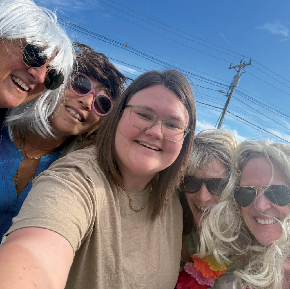 A close-up selfie of five women wearing various blonde and brunette wigs and sunglasses under a clear blue sky.