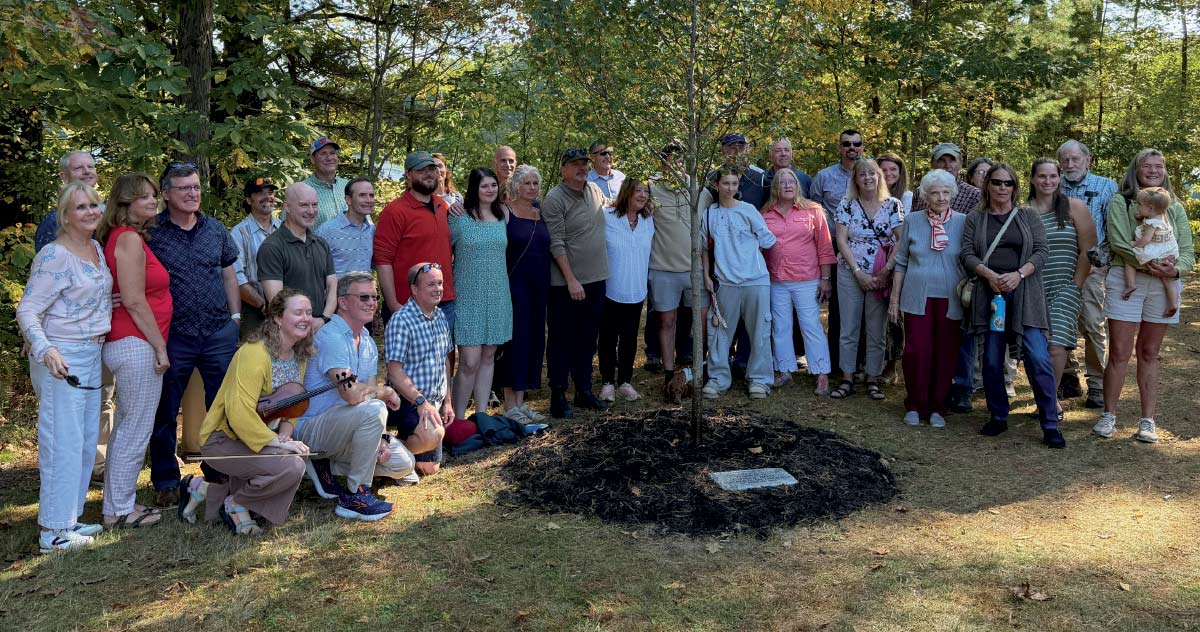 A large group of people gathered in a wooded area for a tree-planting ceremony near a small memorial plaque.