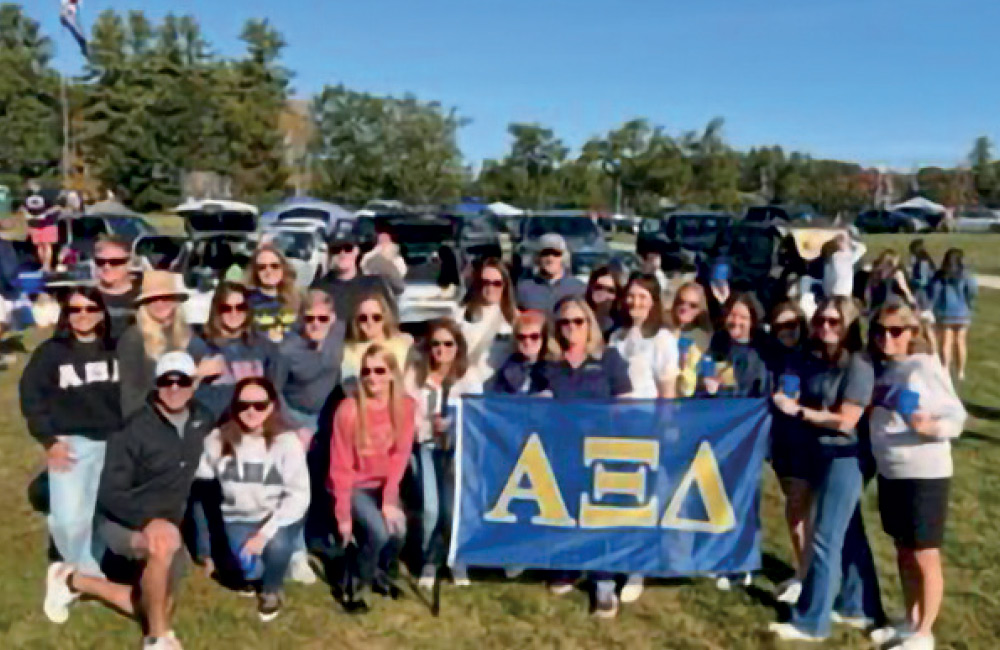 A large group of alumni gathered on a grassy field, posing behind a blue "Alpha Xi Delta" (ΑΞΔ) sorority flag.