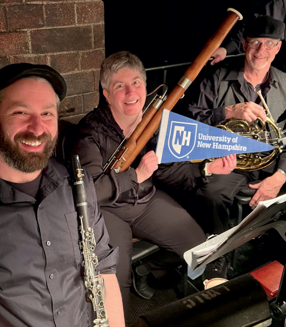 Three musicians in formal black attire smiling while holding a clarinet, a bassoon, and a University of New Hampshire pennant.