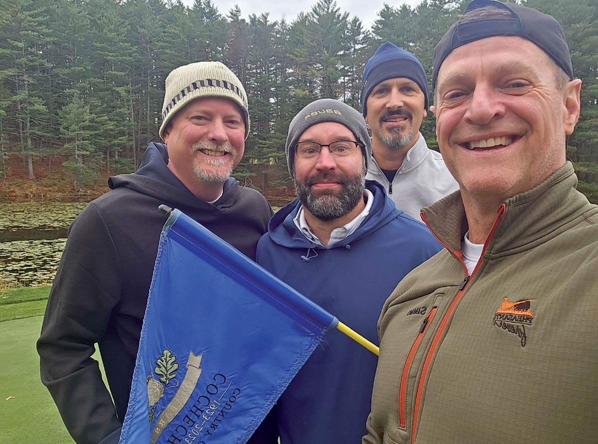 Four men in winter hats and athletic gear posing with a blue "Cochecho Country Club" flag on a golf course.