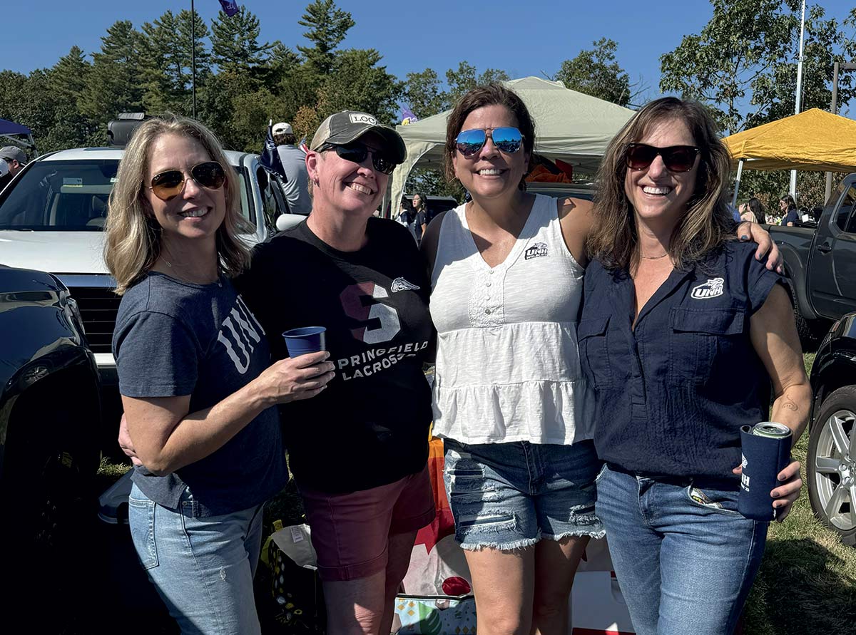 Four women wearing sunglasses and UNH-branded apparel smiling together at an outdoor tailgate event.