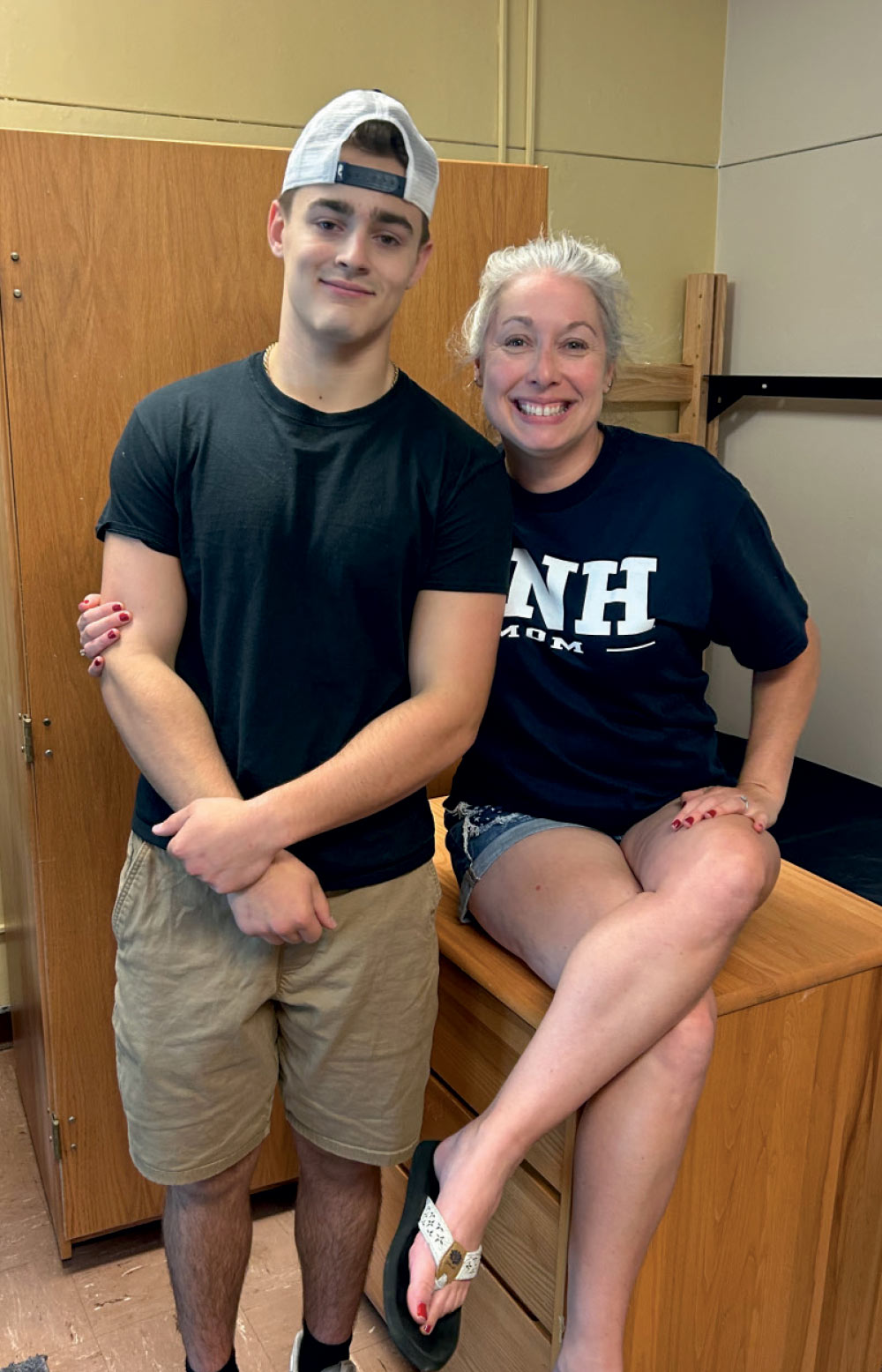 A young man in a black t-shirt and baseball cap standing next to a smiling woman wearing a navy "UNH MOM" shirt.