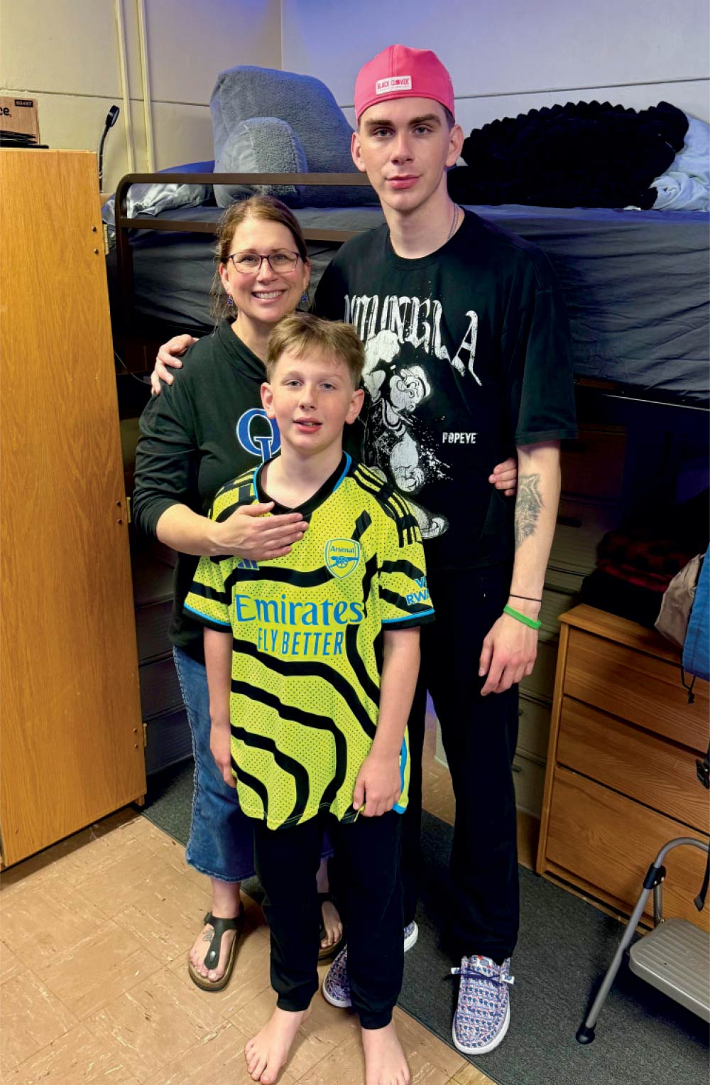 A mother posing with her two sons in a dorm room; the younger son wears a yellow Arsenal soccer jersey.