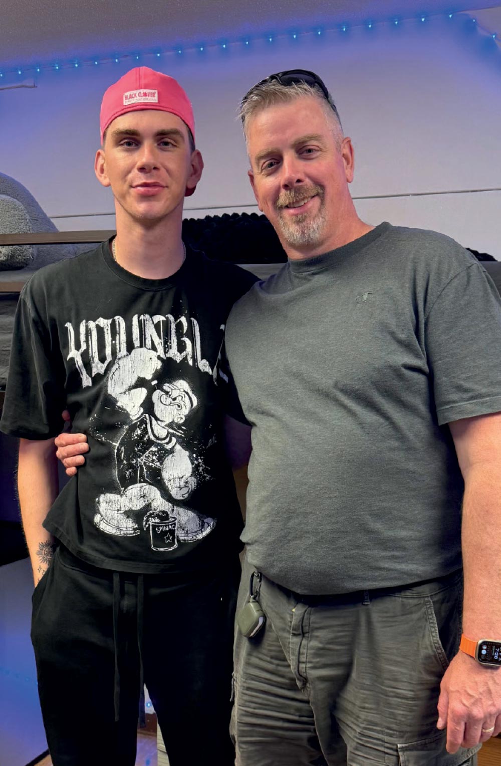 A father and son posing together in a dorm room; the son is wearing a black graphic t-shirt and a pink baseball cap.