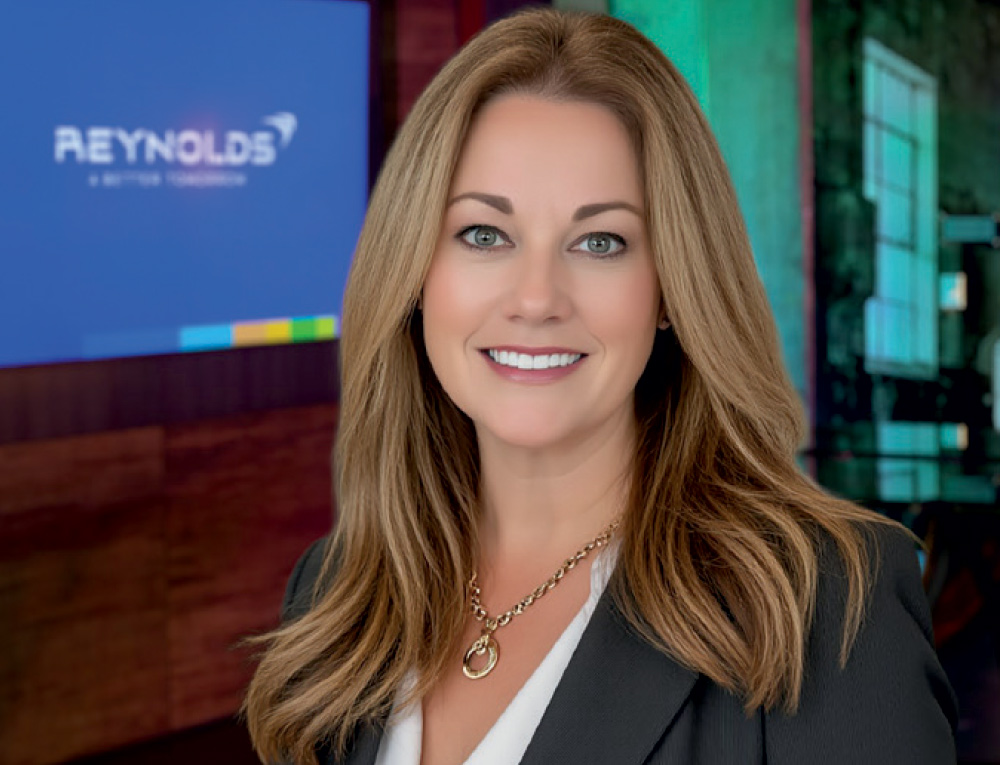 Professional headshot of a smiling woman with long blonde hair, with a blurred "REYNOLDS" logo on a screen in the background.