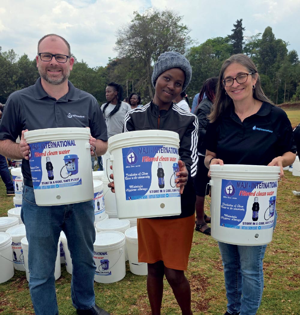 Three people smiling while holding white "Maji International" buckets designed for filtered clean water in an outdoor field.