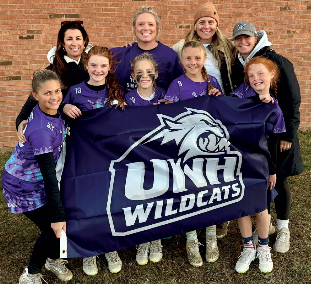 A group of young girls in purple lacrosse jerseys and their coaches posing behind a navy blue UNH Wildcats flag.