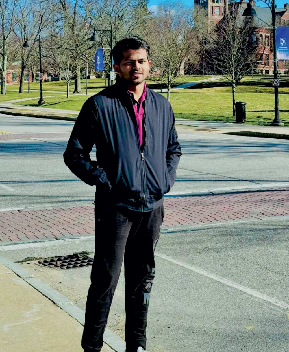 A young man in a dark jacket and purple shirt standing on a sunlit campus sidewalk with brick buildings in the distance.