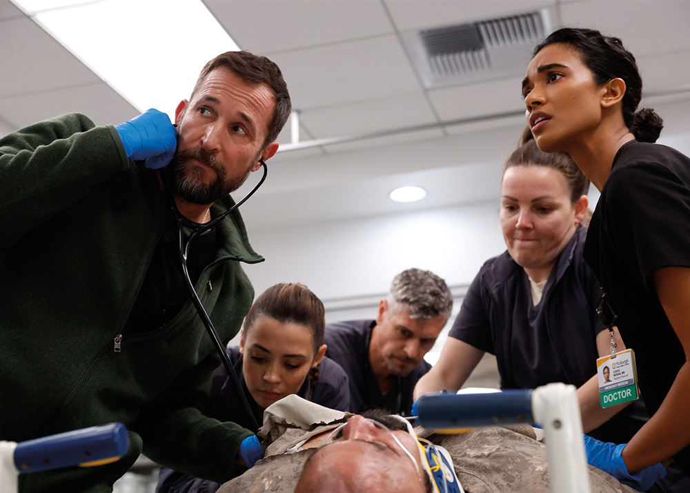 Noah Wyle, using a stethoscope, stands with four medical staff members as they look down at a patient in a neck brace in a scene from “The Pitt.”