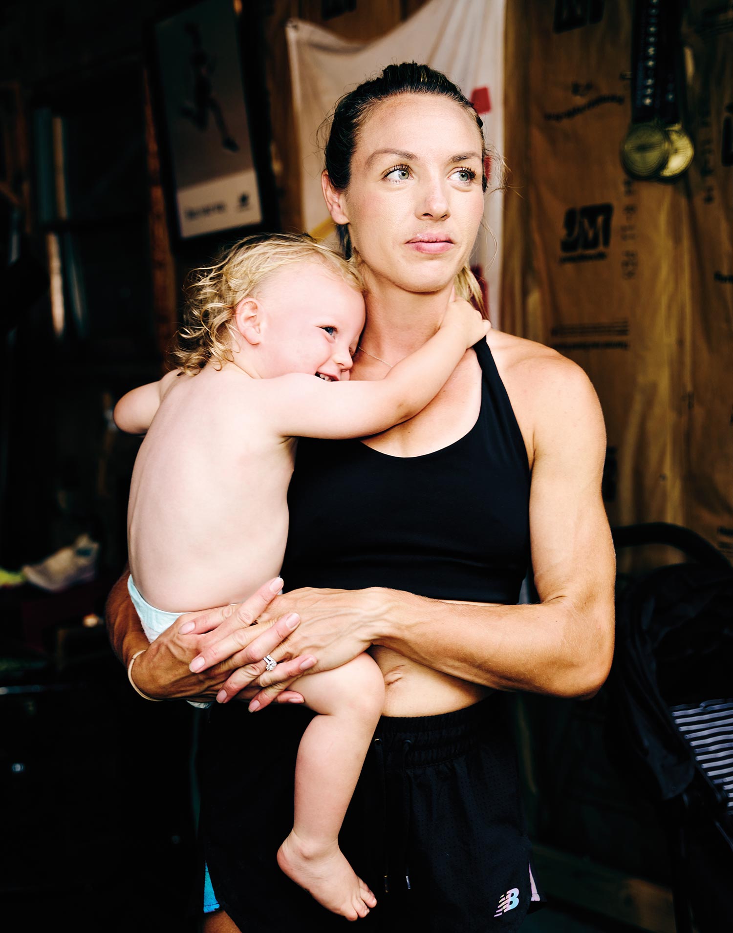 Elle Purrier St. Pierre '18, wearing black athletic gear, holds her young child against her chest. She looks off-camera while standing in a dimly lit indoor space with athletic equipment and medals visible in the background.