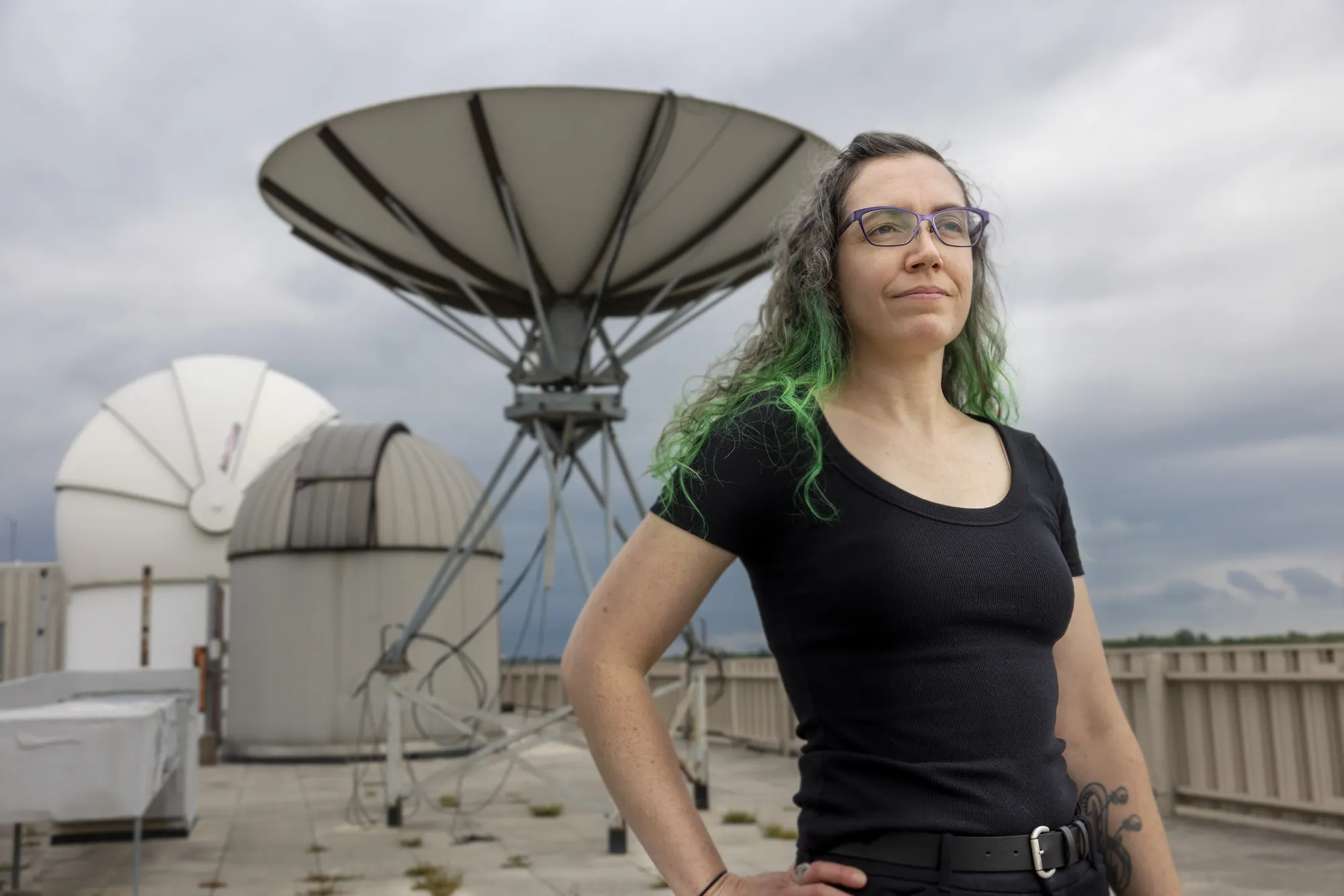 Allison Jaynes stands on a rooftop near large satellite dishes, wearing glasses and a black shirt, with green-highlighted hair visible against a cloudy sky.