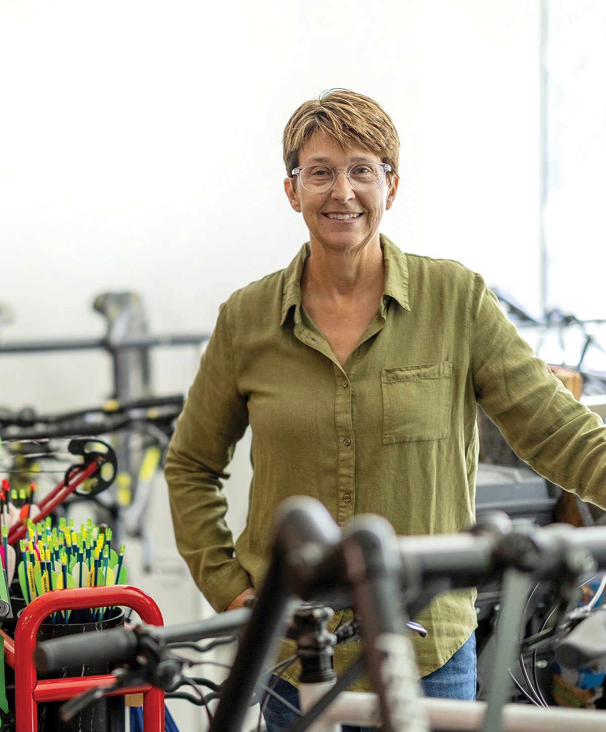 Portrait of a smiling woman with short hair and glasses standing among bicycles and sports gear.