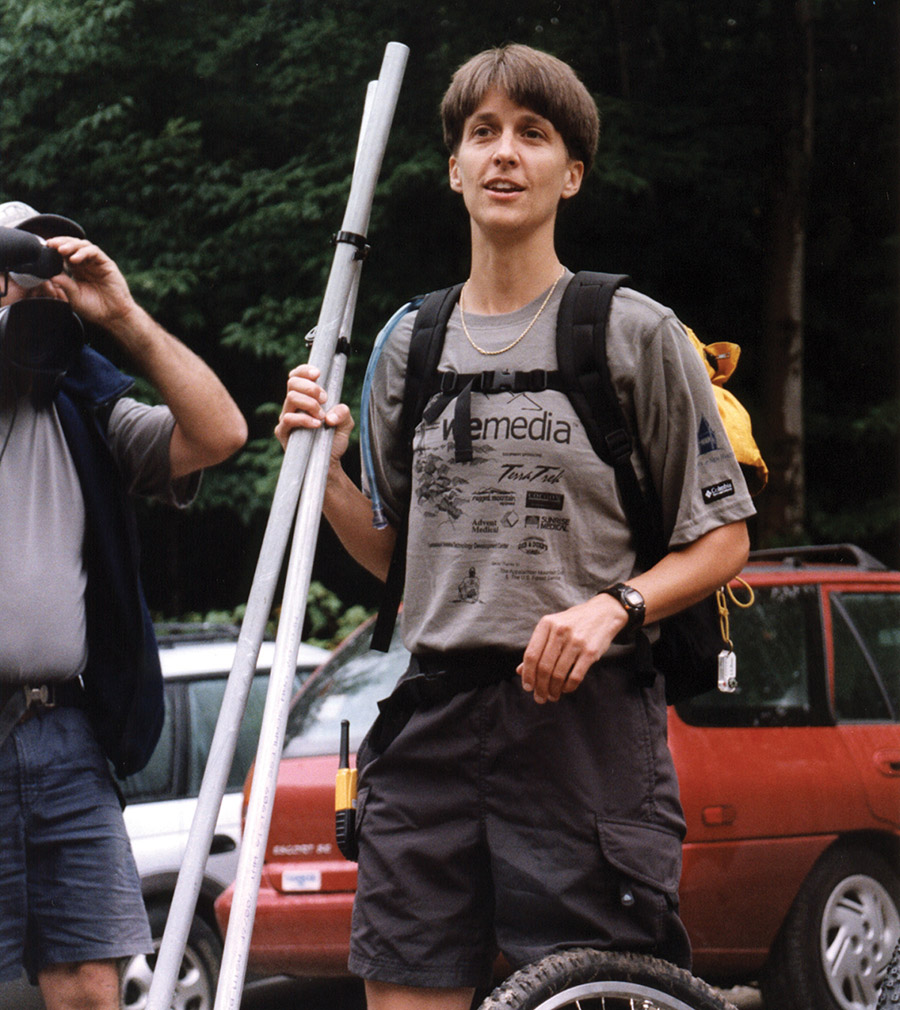 Woman in hiking clothes and a backpack carrying long metal poles outdoors near vehicles.