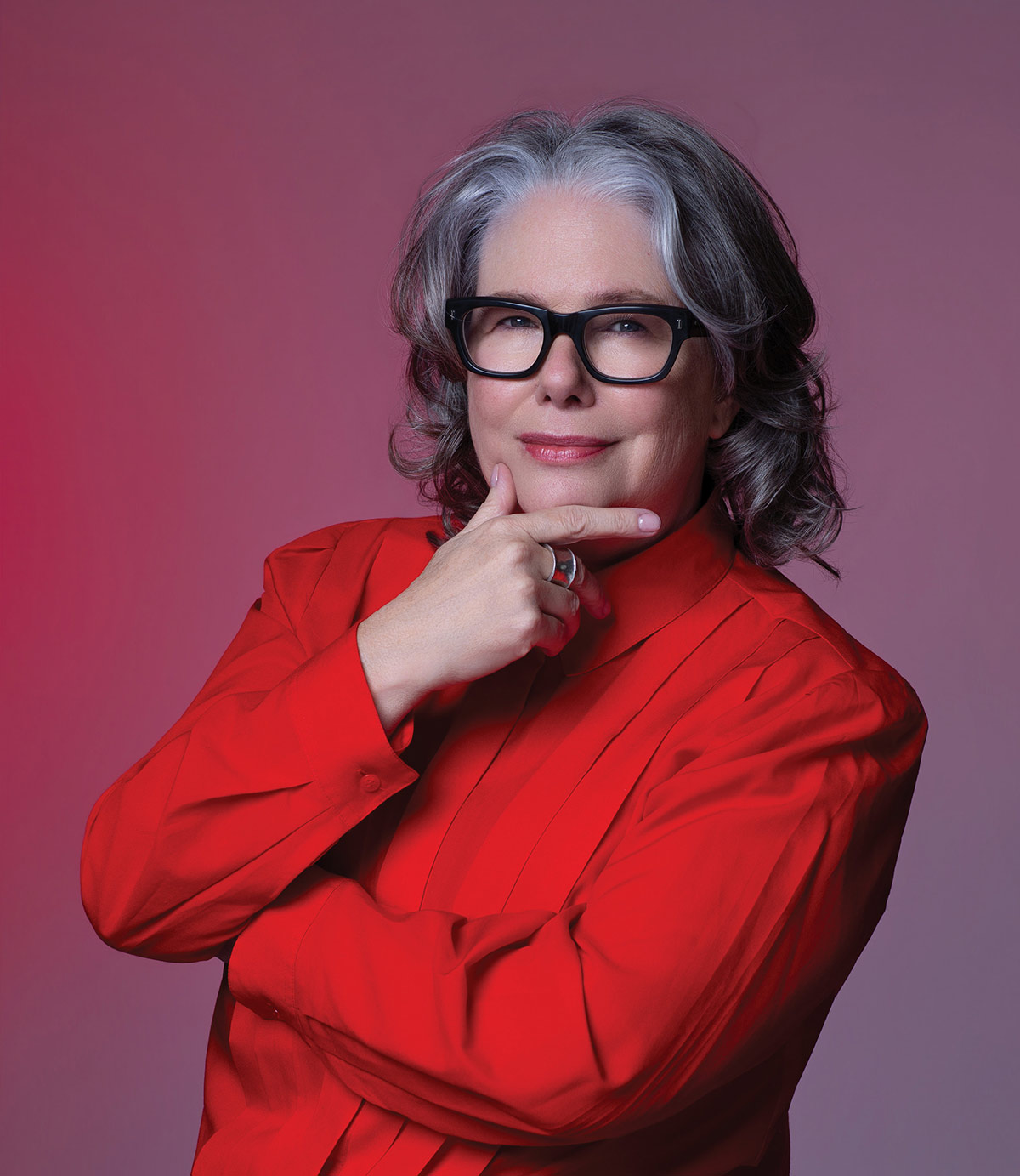 Woman with grey hair and glasses posing thoughtfully in a red shirt.