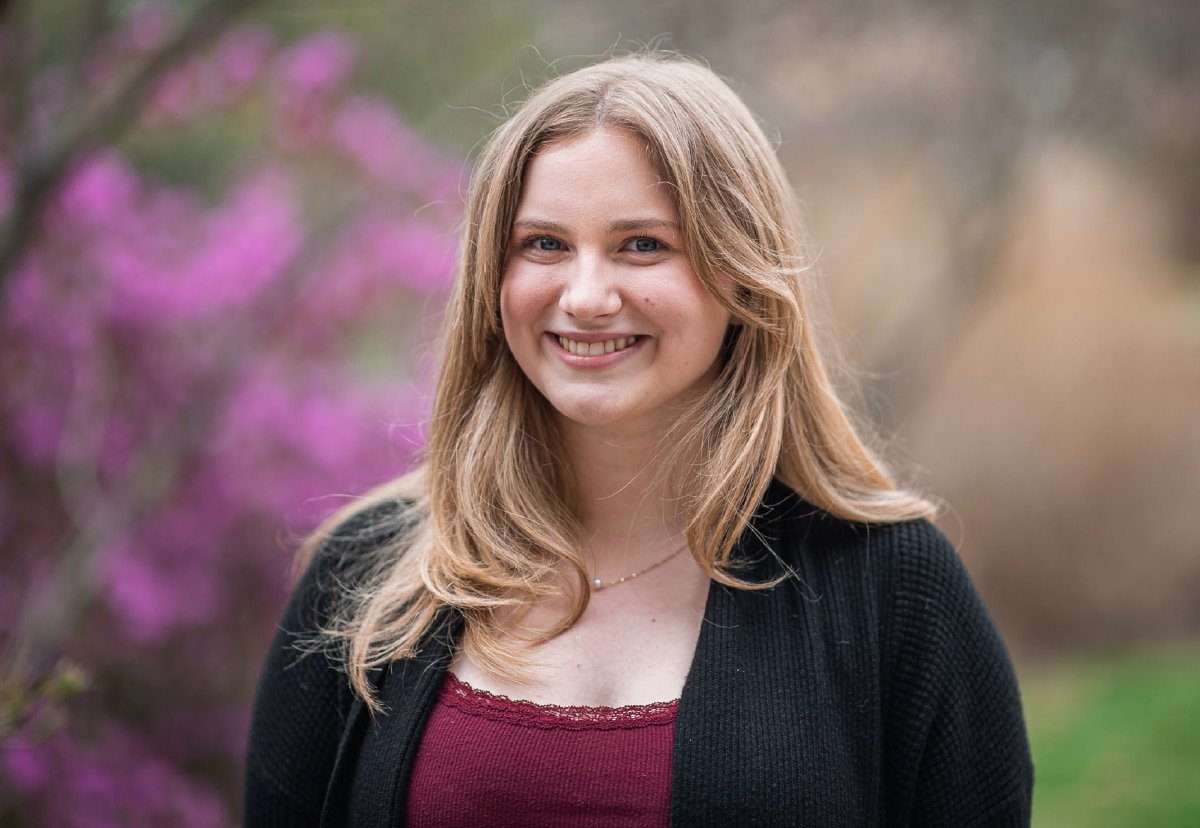 Smiling blonde woman in a black cardigan outdoors in front of blurry pink flowers.