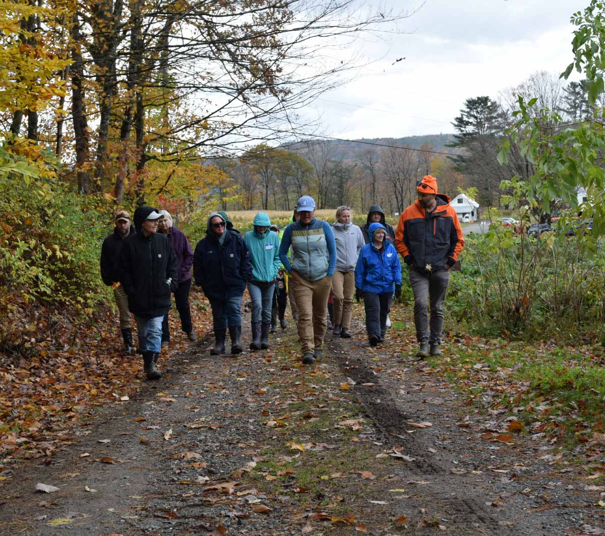 Group of people in jackets walking along a dirt path lined with autumn trees and fallen leaves.