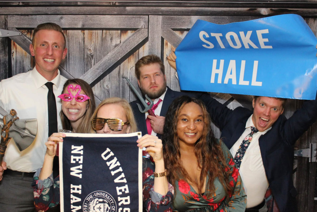 Group of friends posing in photo booth props holding a "Stoke Hall" banner and a University of New Hampshire pennant.