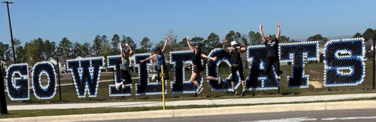Five people jumping in the air in front of a chain-link fence spelling out "GO WILDCATS".