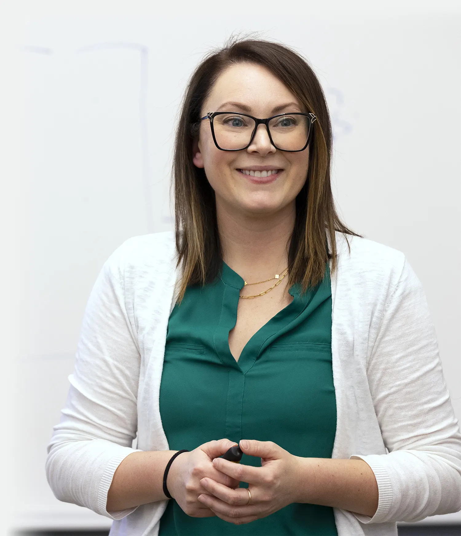 A smiling woman wearing black-framed glasses, a green blouse, and a white cardigan, holding a marker in front of a whiteboard.