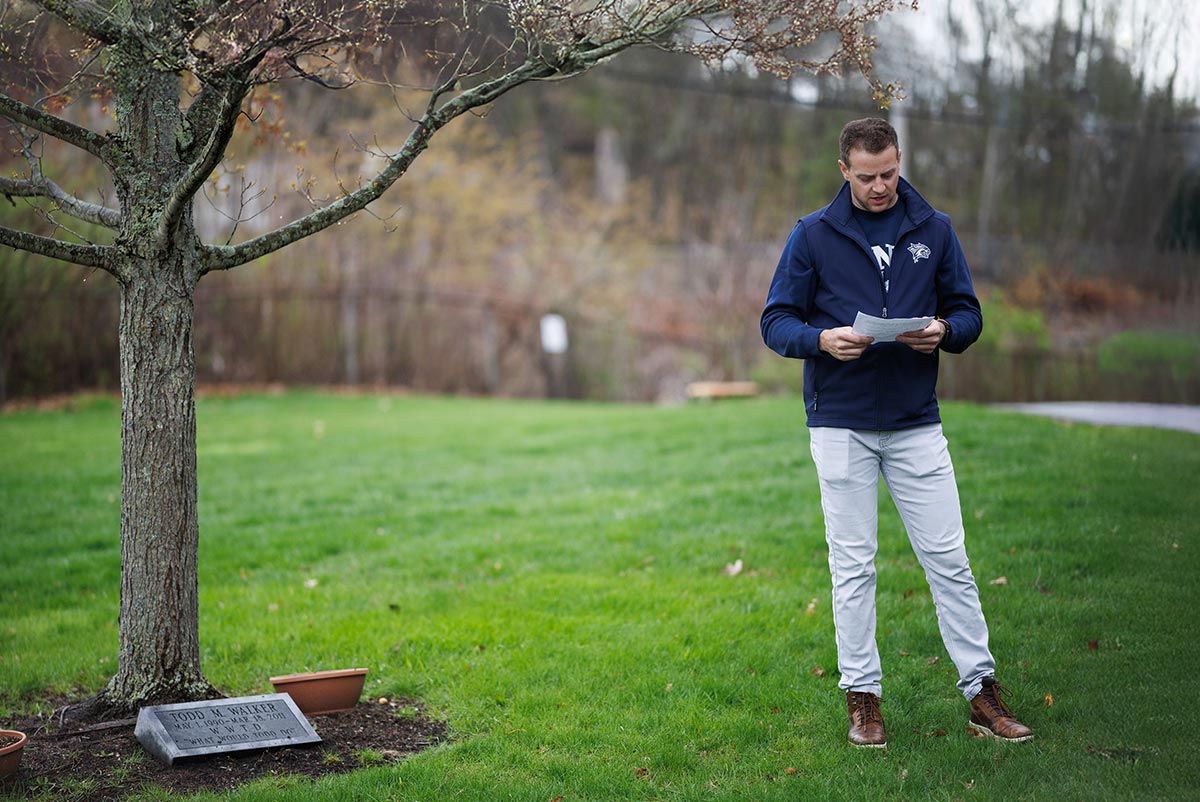 A man in a navy UNH jacket reads from a paper beside a tree with a memorial plaque dedicated to Todd M. Walker.