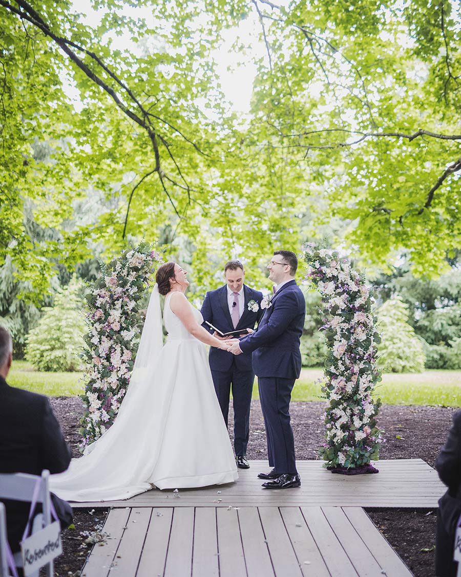 A bride and groom laughing together during an outdoor wedding ceremony framed by floral arches.