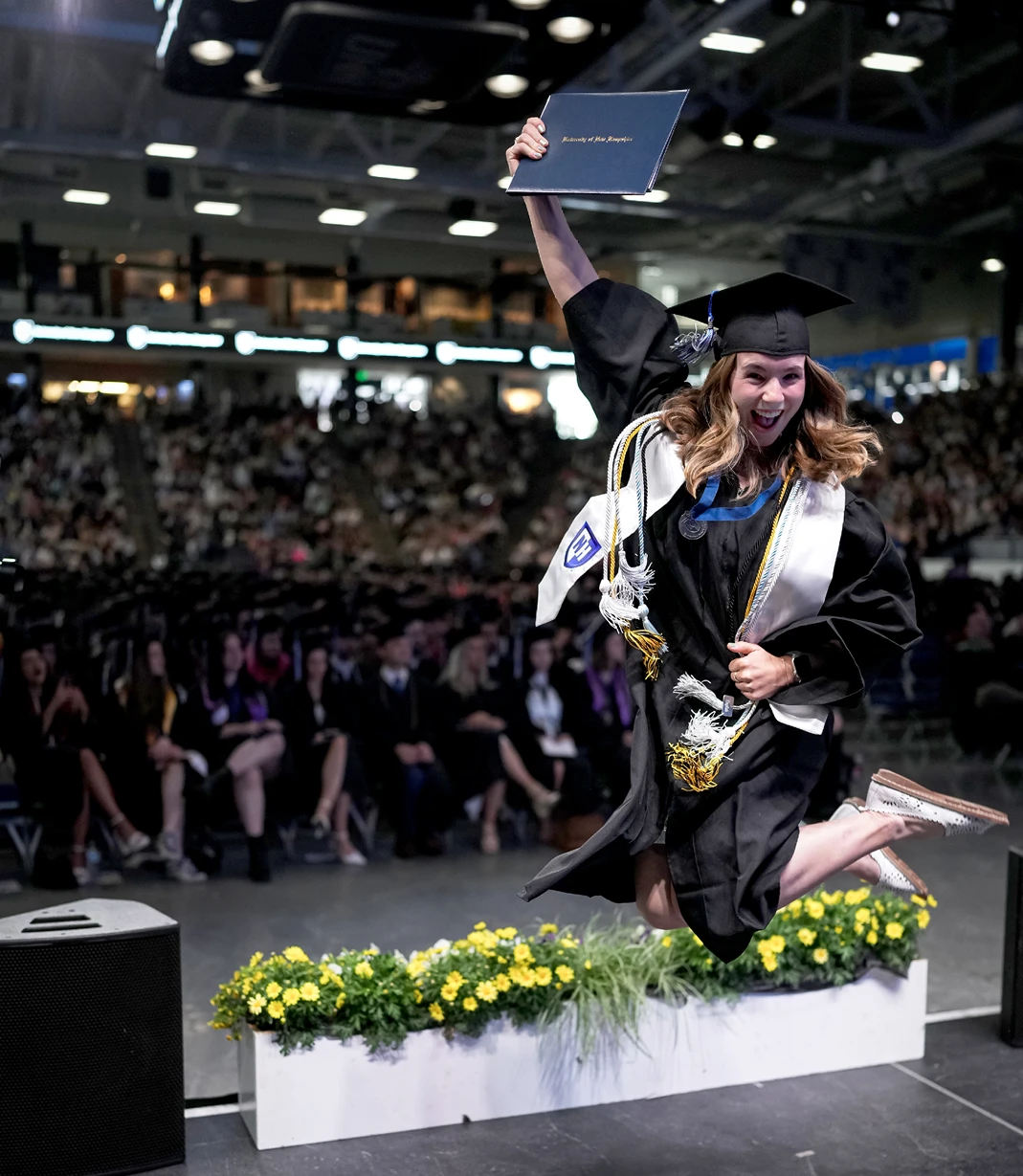 A University of New Hampshire graduate jumps in celebration while raising her diploma before the commencement audience.
