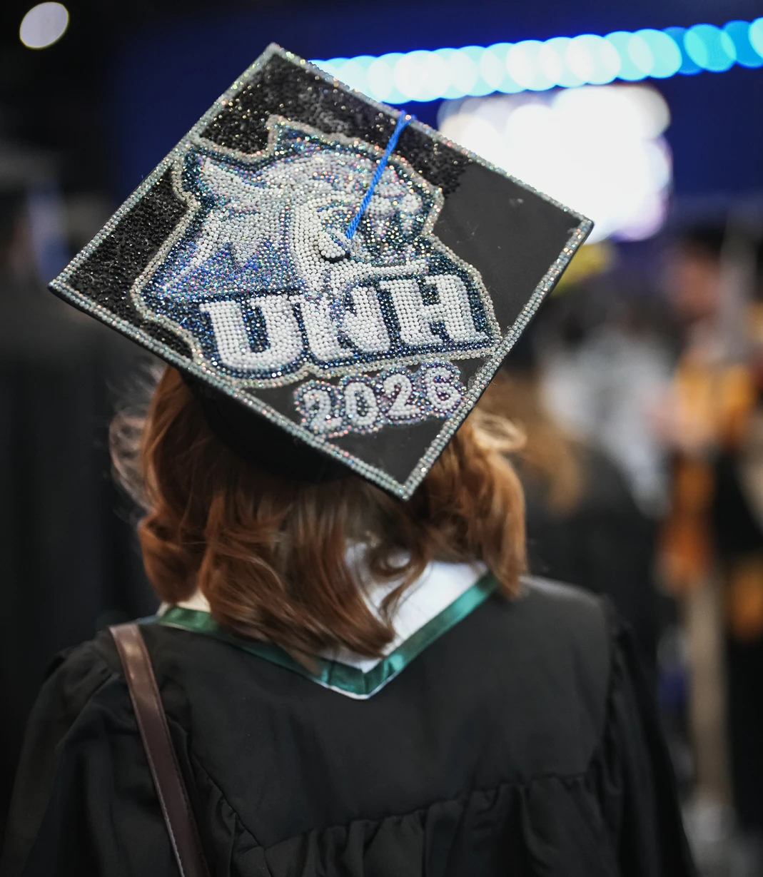 A graduate wears a rhinestone-covered mortarboard decorated with the UNH Wildcat logo and “2026.”