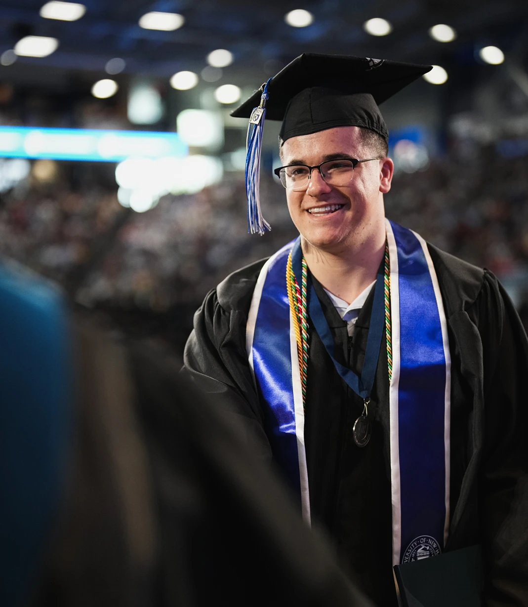 A University of New Hampshire graduate wearing glasses, honor cords, a medal, and a blue stole smiles during commencement.
