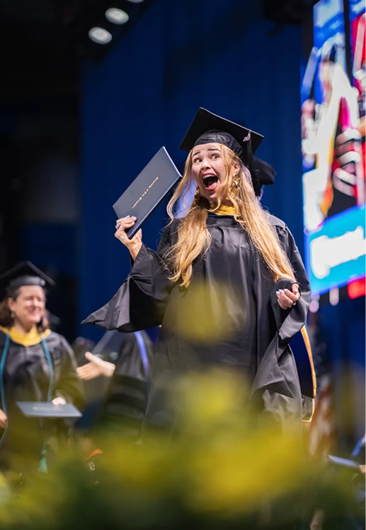 A graduate celebrates onstage while holding up her diploma during the University of New Hampshire commencement ceremony.