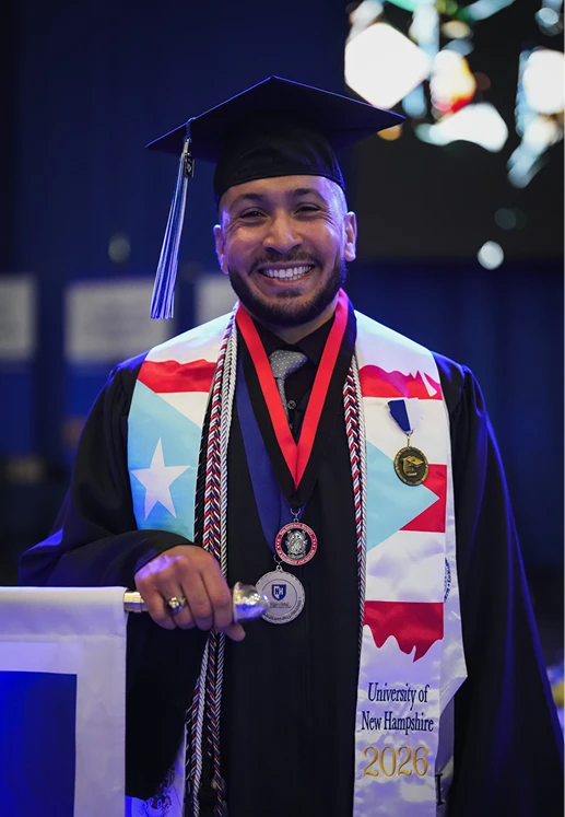 A smiling University of New Hampshire graduate wears medals, honor cords, and a Class of 2026 stole featuring the Puerto Rican flag.
