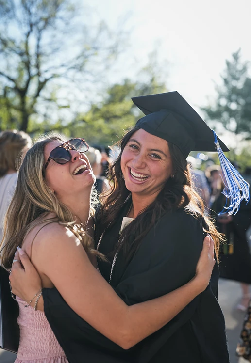 A University of New Hampshire graduate and another woman laugh while embracing outdoors after commencement.