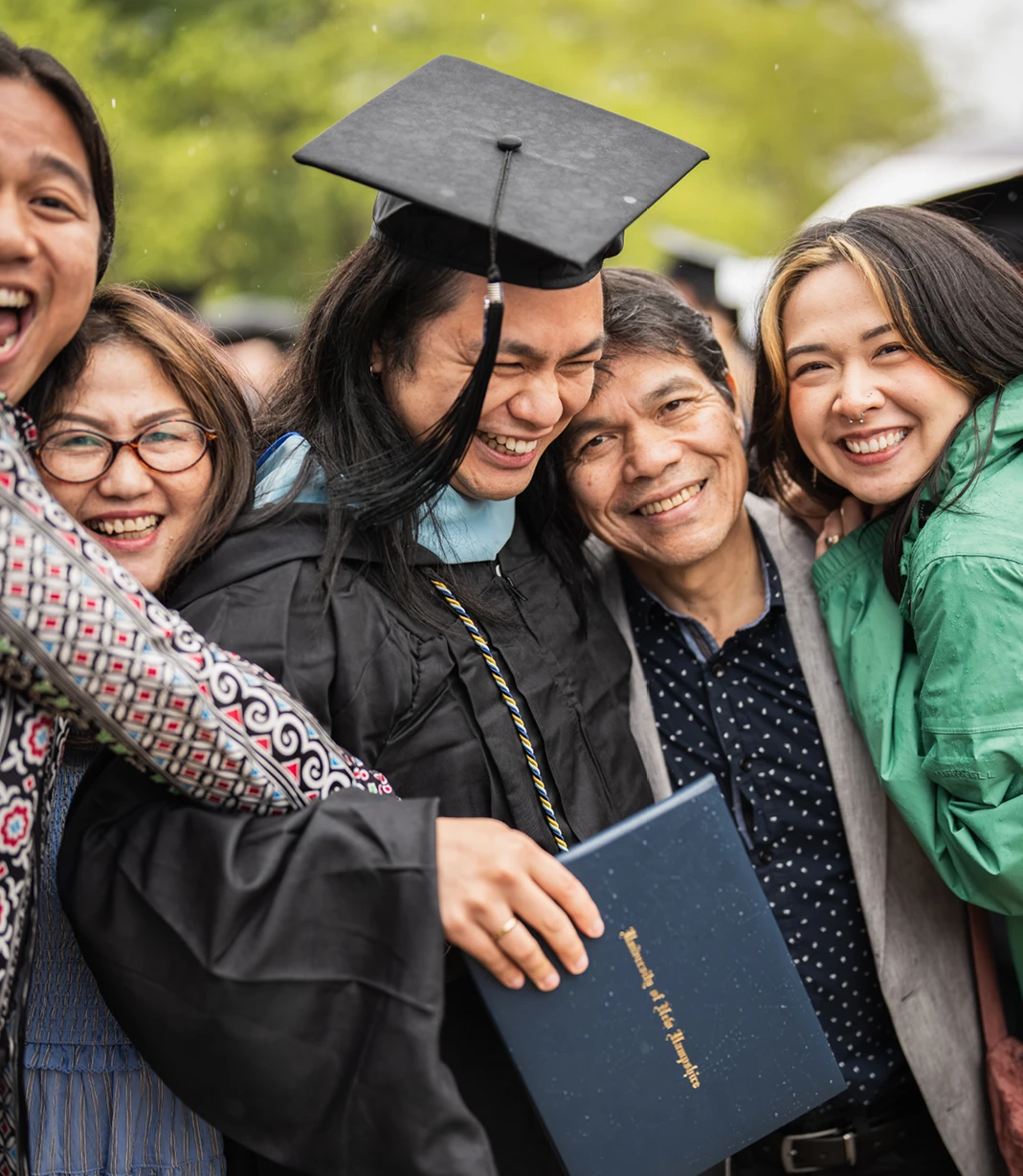 A University of New Hampshire graduate holds a diploma and laughs with family members gathered around them in the rain.
