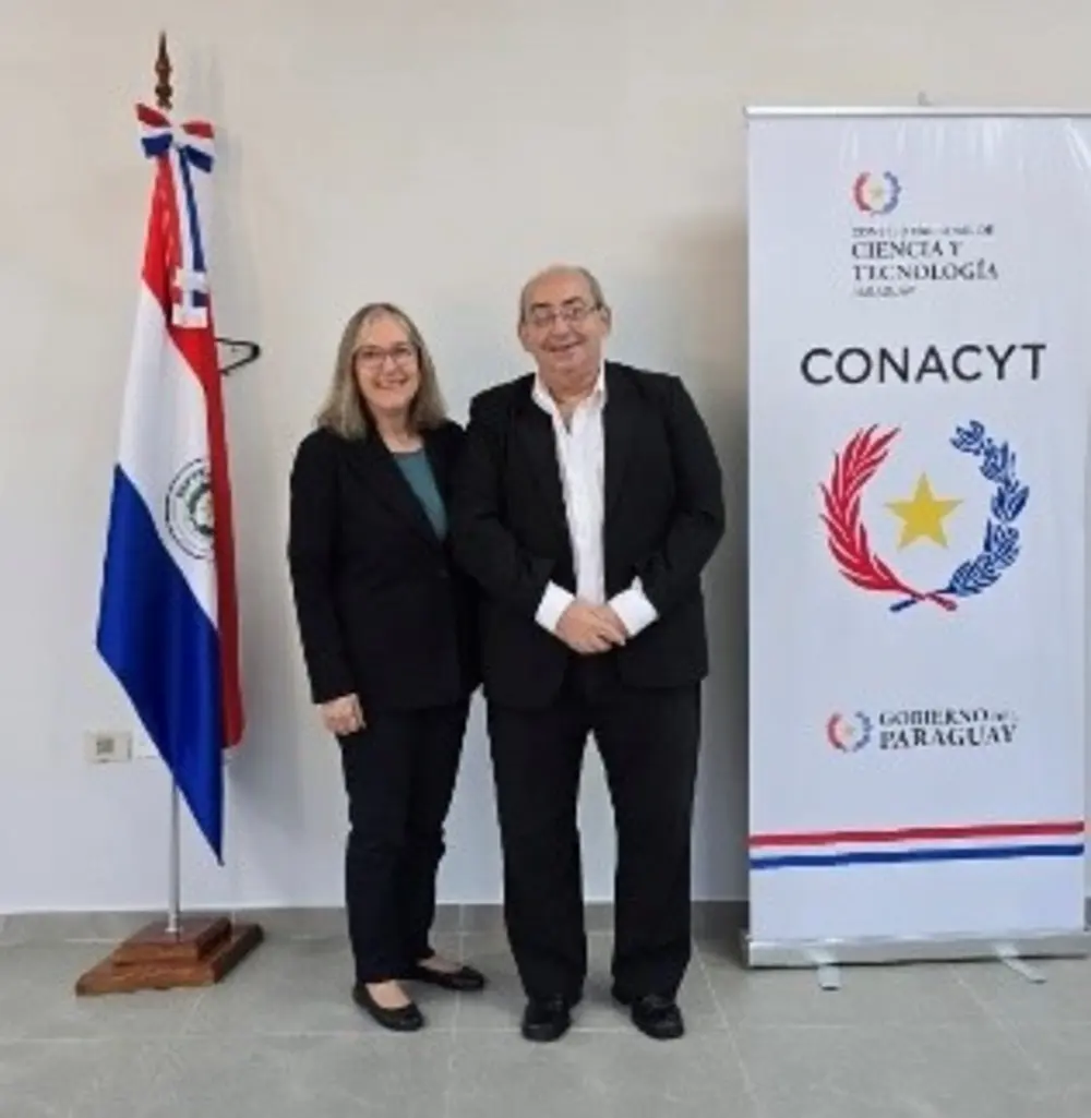 Denise Dumas LeBlanc ’74 stands with D.Sc. Benjamin Barán beside the Paraguayan flag and a CONACYT banner.