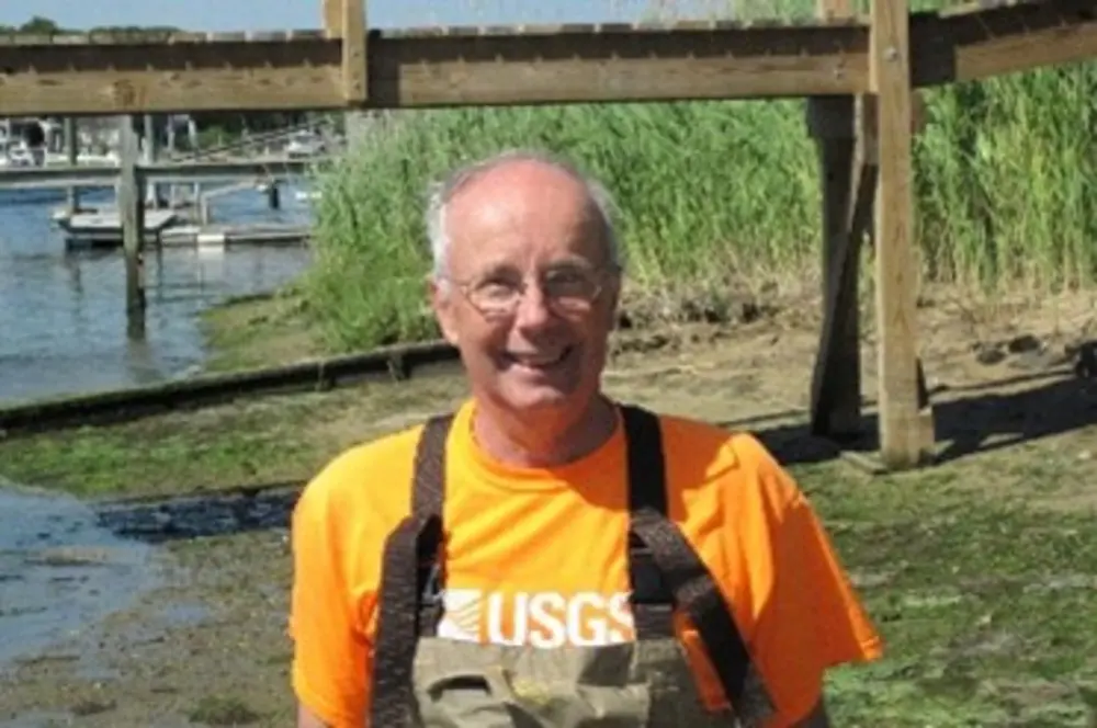 Denis LeBlanc ’75 smiles outdoors while wearing an orange USGS shirt and chest waders.