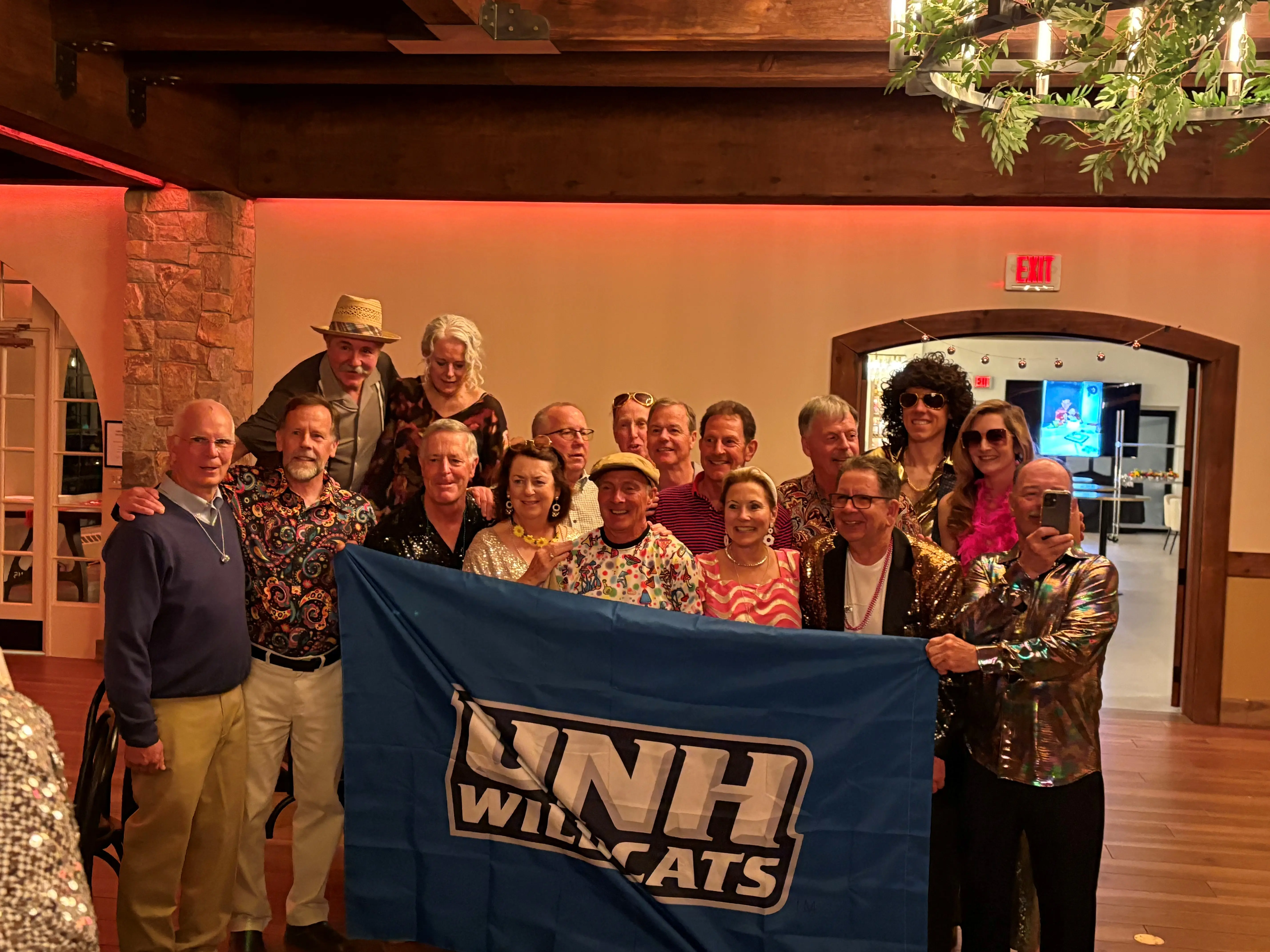 A group of UNH alumni and friends dressed in 1970s-themed attire holds a UNH Wildcats banner at a birthday gathering.