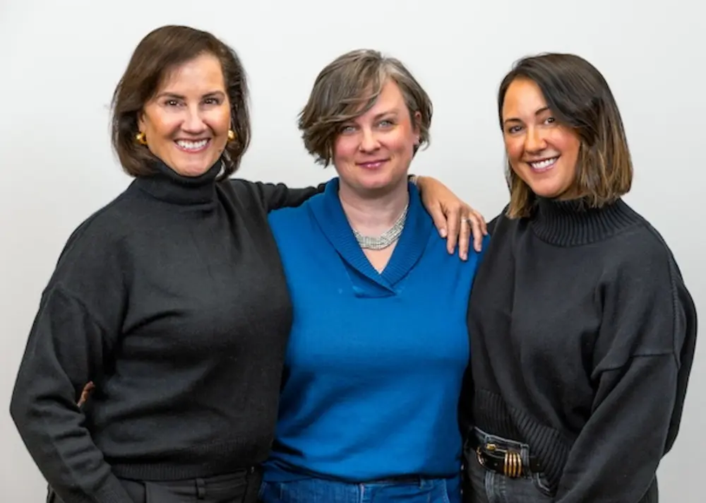Three women stand together for a portrait against a light-gray background.