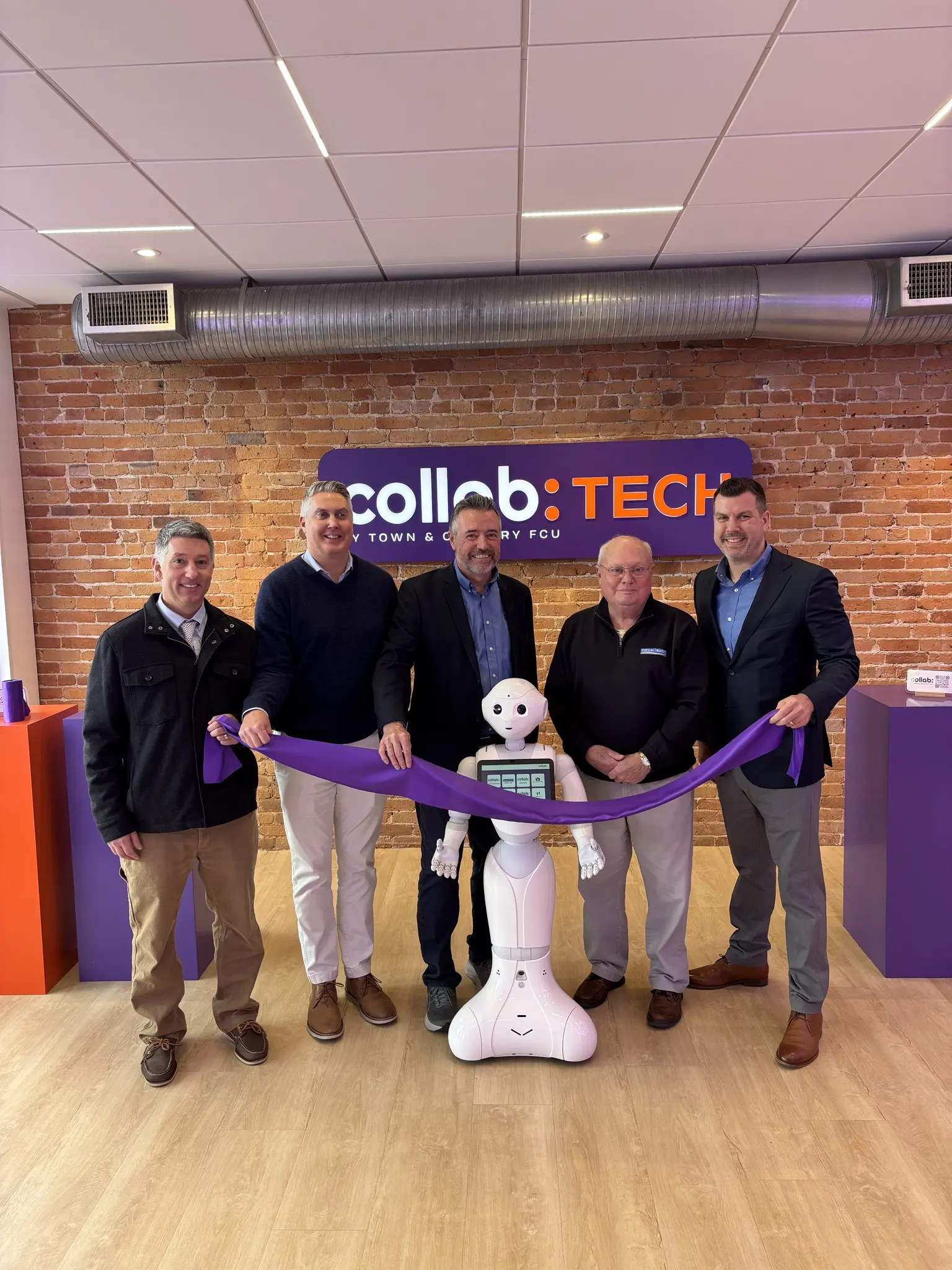 Ross Elliott ’99, Josh Fifield ’99 and other Town & Country Federal Credit Union officials cut a purple ribbon at a new Portland workspace as a white service robot stands nearby.