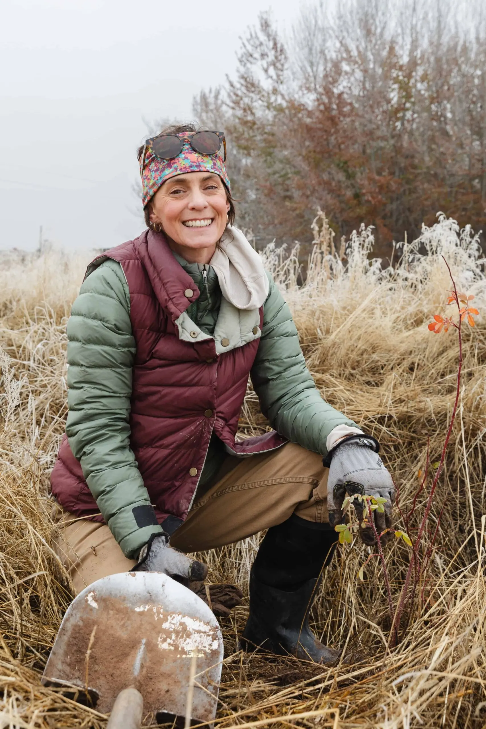 Janice Gardner ’04 kneels in a grassy field while wearing outdoor gear and smiling beside a covered bird used in falconry.