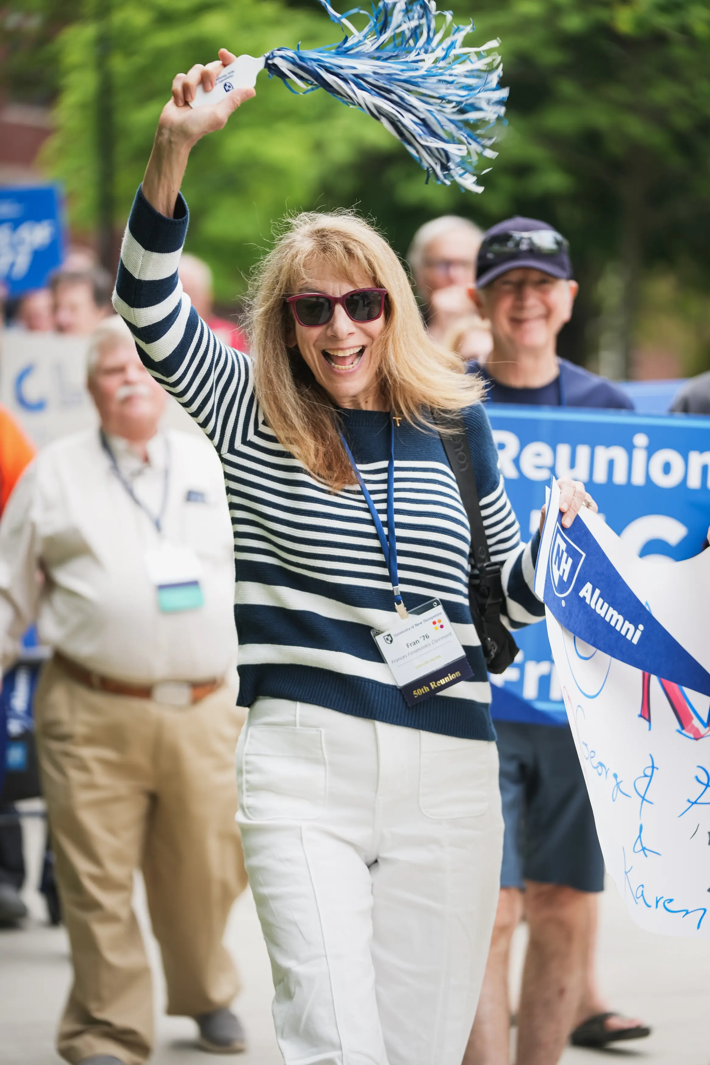 A smiling UNH alumna in sunglasses waves a blue-and-white pom-pom while walking in a reunion parade.