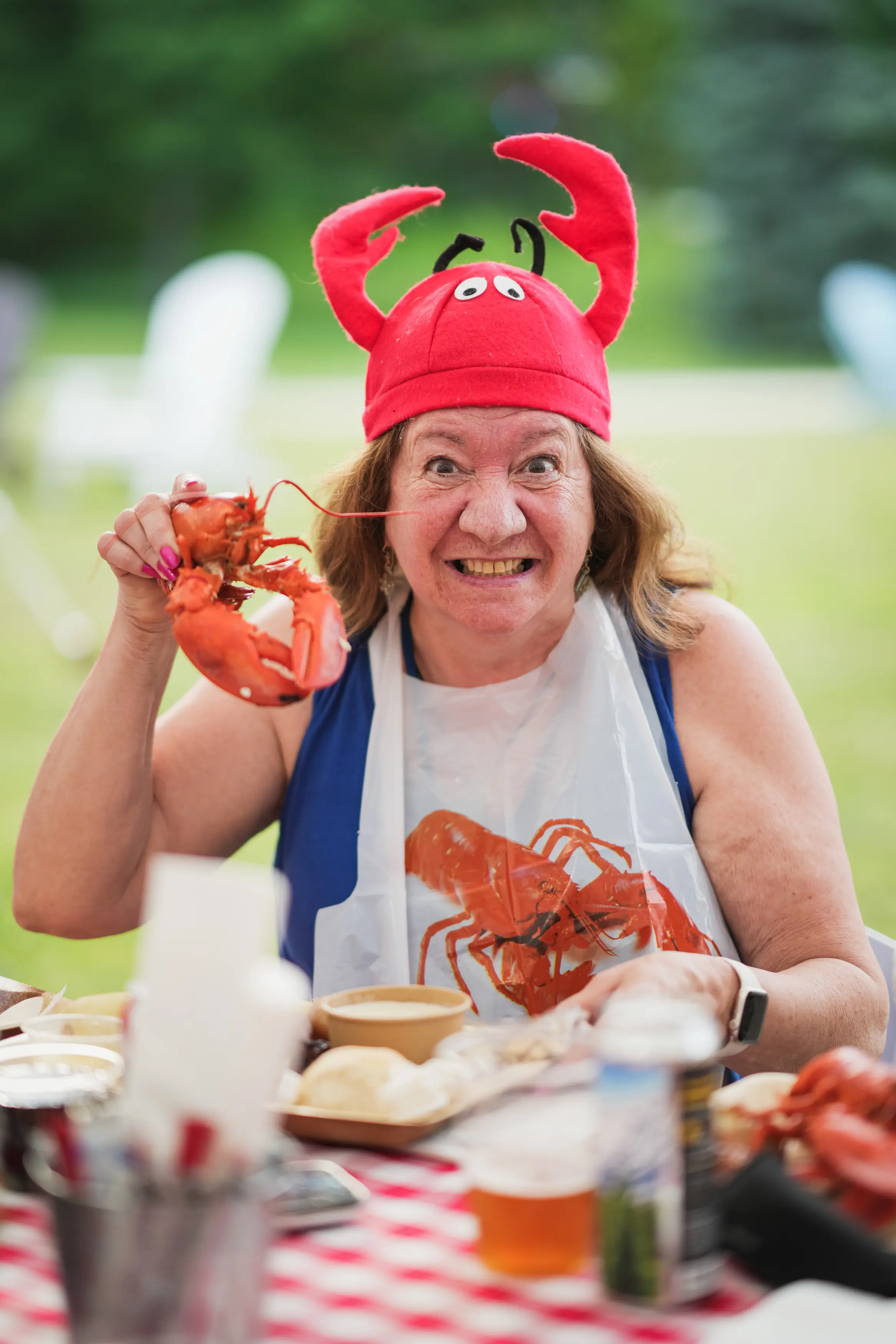 A smiling alumna wearing a red lobster hat and lobster bib holds up a cooked lobster at an outdoor reunion meal.