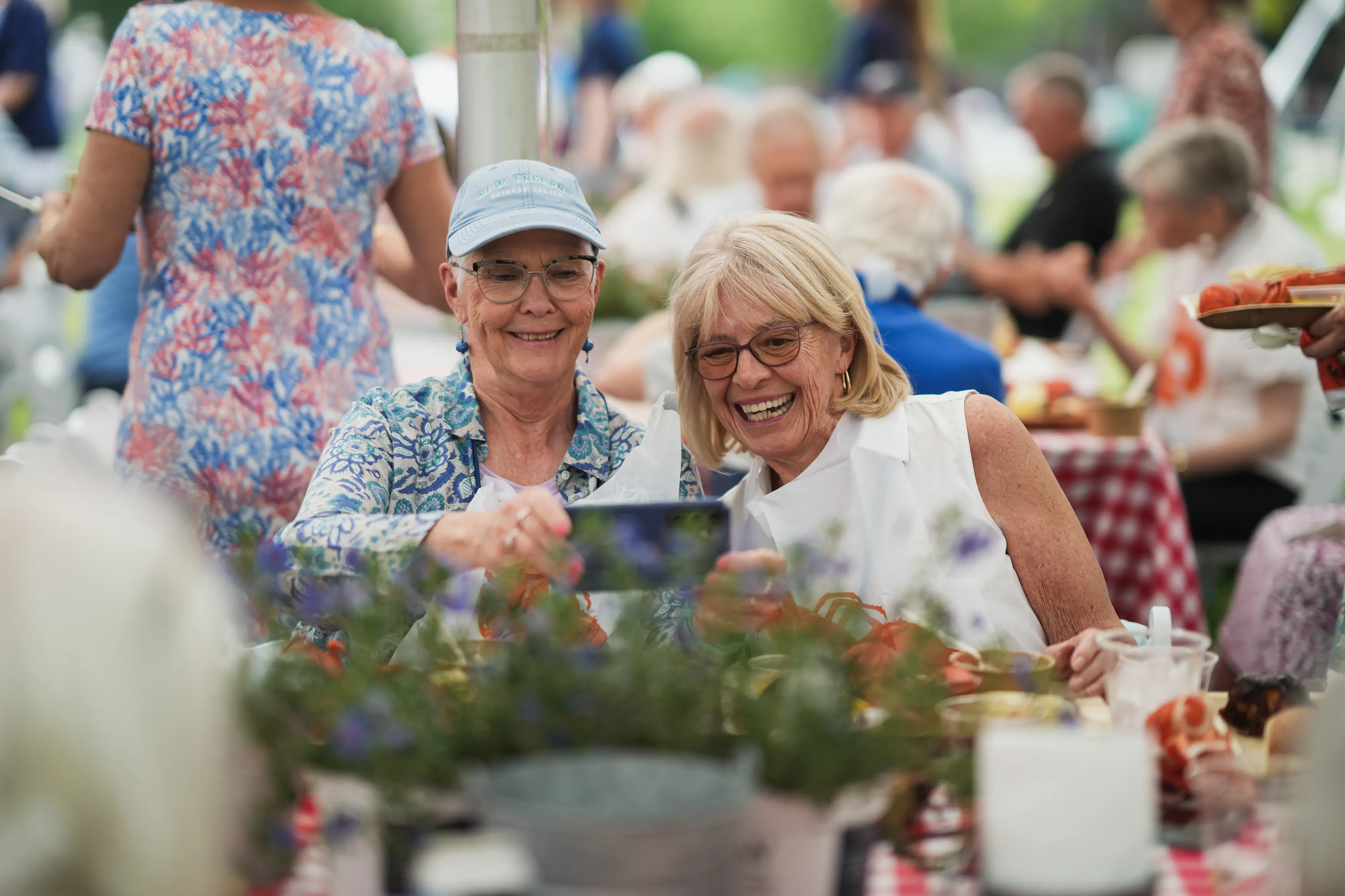 Two alumnae smile over a phone while seated together at an outdoor lobster luncheon.