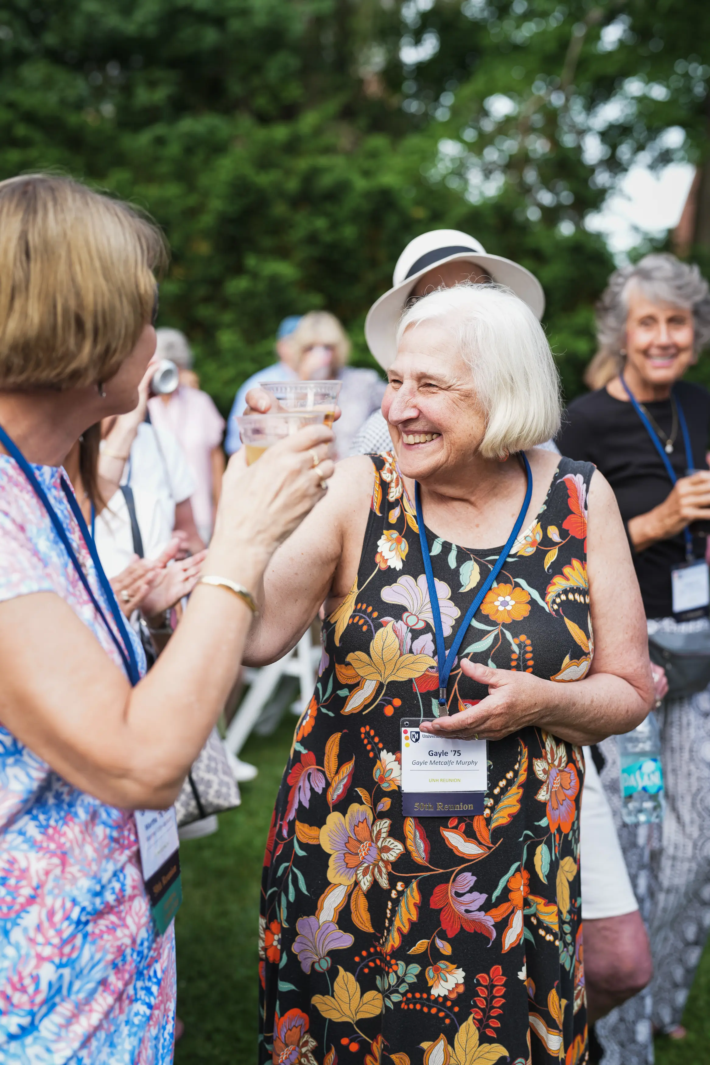 Two alumnae raise plastic cups in a cheerful toast during an outdoor reunion gathering.