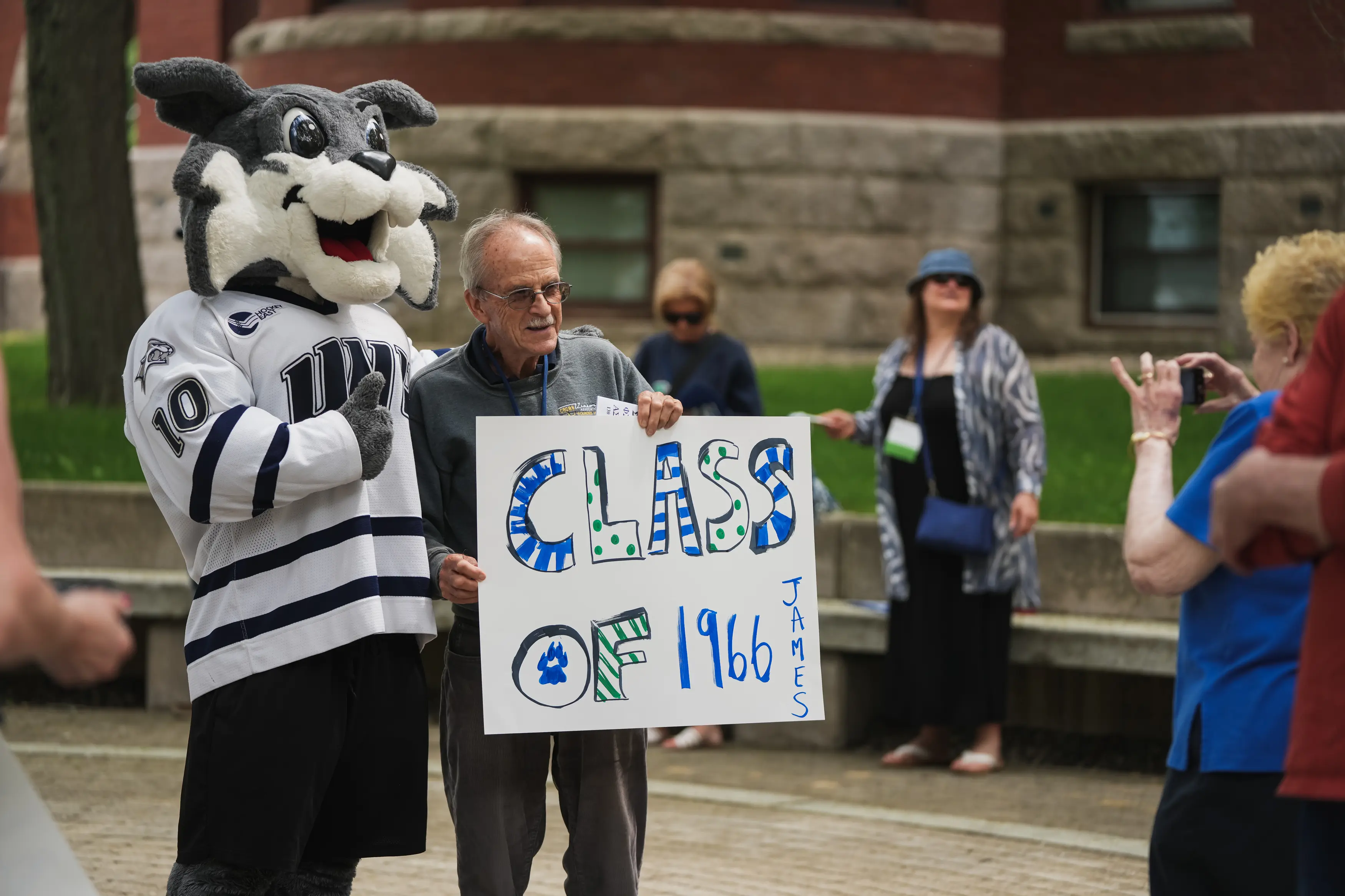 Wild E. Cat poses beside an alumnus holding a handmade “Class of 1966” sign as another attendee takes their picture.